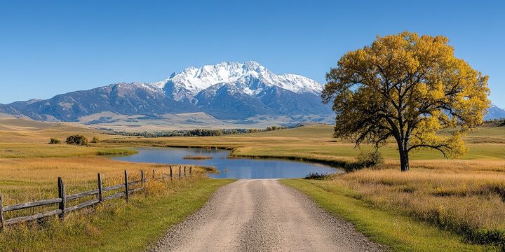 Journey to Majesty: An open dirt road stretches towards the majestic snow-capped mountains under a vast clear sky, and a lone tree in golden foliage next to a lake in landscape. - Powered by Adobe