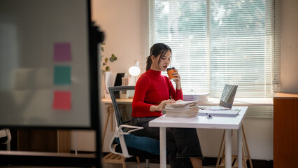 Professional Asian woman reviewing paperwork while sipping coffee at workplace
