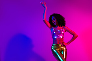 Stylish young woman dancing in vibrant neon lighting with glowing purple and pink tones, wearing modern metallic fashion