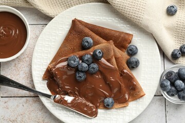 Delicious chocolate crepes with sweet paste and blueberries on light tiled table, flat lay