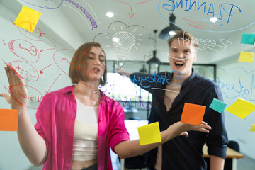 Couple of businesspeople discussing and brainstorming about solving marketing problem in front of glass board at business meeting by using mind map and sticky notes. Working together. Immaculate.