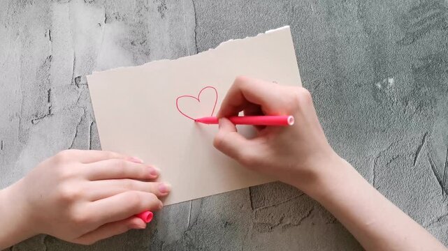 young woman draws a heart on a piece of paper and then roughly crumples it in her hands. A top view of female hands against a gray concrete table