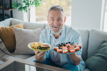 Smiling Elderly Man Enjoying a Meal with Plates of Sushi and Burger with Fries at Home in a Bright Living Room