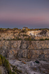 A rocky cliff with sparse trees and dry bushes under warm sunlight. At the top, an abandoned industrial structure stands among autumn foliage, creating a contrast between nature and human-made ruins.