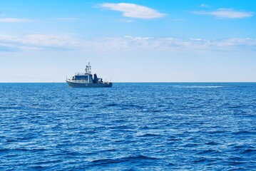 The boat is in the center of the photo, and the water is a deep blue color. The sky is clear and blue with white clouds on the horizon, and the waves are small and gentle.