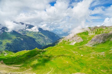 Fototapeta premium A stunning landscape features green rolling hills and distant snow-capped mountains. The blue sky is dotted with white clouds, creating a serene and tranquil atmosphere.