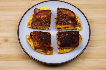 A homemade breakfast sandwich cut in quarter with crispy toasted bread, scrambled eggs, bacon, and melted cheese on a plate over a wooden surface. A rustic, hearty meal perfect for breakfast or brunch