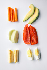 Assorted fresh food ingredients arranged on a white background, including carrot sticks, avocado slices, apple pieces, red bell pepper, banana chunks, and boiled egg halves
