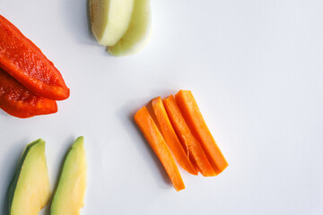 Assorted fresh food ingredients arranged on a white background, including carrot sticks, avocado slices, apple pieces, red bell pepper, banana chunks, and boiled egg halves
