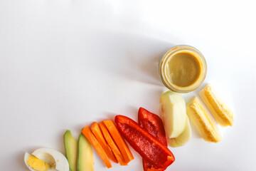 A glass jar of homemade baby puree with fresh vegetables and fruits arranged on a white background. Includes carrot, bell pepper, apple, and banana. Healthy organic food for infants and toddlers
