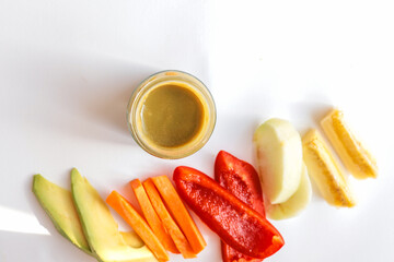 A glass jar of homemade baby puree with fresh vegetables and fruits arranged on a white background....