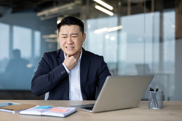 Fototapeta premium Asian young man businessman sits in the office at the table and holds his hand on his neck, feels a strong pain in his throat