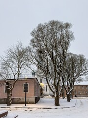 A quiet winter street scene featuring snow-covered trees, a small wooden house, and a distant church with blue domes. The cold atmosphere and evening light create a peaceful and nostalgic setting