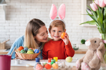 Naklejka premium Little nice boy showing painted egg for Easter with his young mom at home. Small kid child son making springtime holiday decorations together with young mother in the kitchen