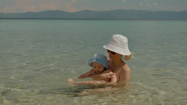 A mother relaxes in the calm sea, cradling her baby while the gentle waves lap against them. They share a peaceful moment together on a summer vacation.