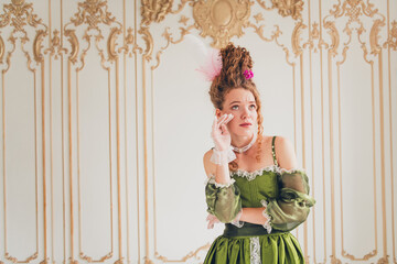 Elegant young woman in historic green dress posing indoors with baroque decor, emphasizing a classic and vintage comparison of past and modern life themes.