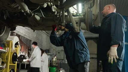 Car engineer holds light while assisting colleague working under vehicle, adjusting hand position to point out something specific in car mechanism