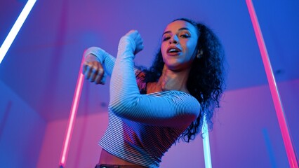 Low angle camera of hispanic dancer looking at camera and dancing to city pop music. Professional attractive hipster moving to hip hop song with purple led or neon light. Stylish cloth. Regalement.
