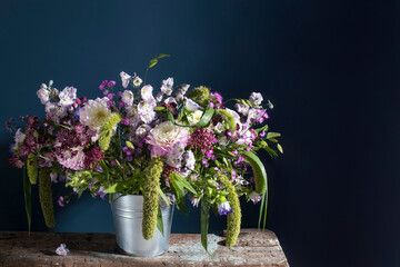 A bouquet of delicate pink dahlias, Garden delphiniums, Setaria macrocheata, astrantias, and woodland bluebells on a rough, unpainted bench surface against a dark blue background. Space for text.