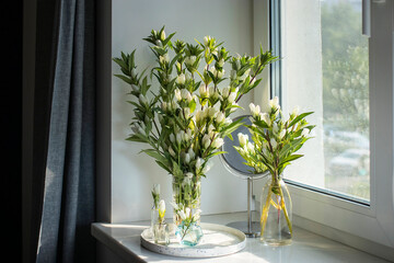 Several bouquets of Gentiana White Sky GENTI WHITE SKY for interior decoration placed on a windowsill with a round mirror next to gray curtains.