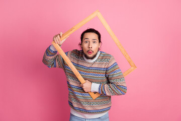 Young man with surprised expression holding a wooden frame against pink background while dressed in a stylish patterned winter sweater