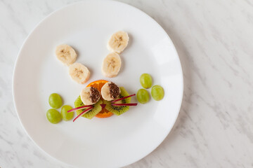Creative healthy snack of breakfast for kids fruit bunny shaped on white plate, easter eggs around. Rabbit, basket makes from grape, orange, banana, apple. Top view