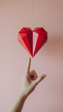 A delicate hand reaching toward a geometric red heart suspended in mid-air against a soft pink background ai generated