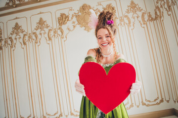 Elegant young woman in baroque costume holding a red heart with a smile in a decorated castle...