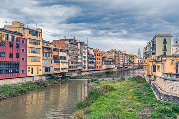 Colorful houses line the Onyar River in Girona, Spain, reflecting in the water under a cloudy sky. Perfect for travel, architecture, and cityscape themes