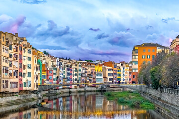 Fototapeta premium Scenic view of colorful houses along Onyar River and Puente Gomez bridge in Girona during sunset, Spain. Beautiful reflection of historical buildings in calm water creates magical atmosphere