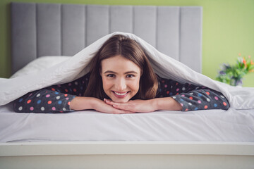 Young smiling woman lying under a blanket in bed wearing polka-dot pajamas, exuding happiness and...