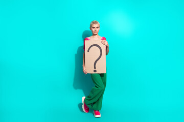 Stylish young woman in casual outfit holding a cardboard sign with a question mark on vivid turquoise background