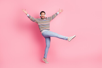 Joyful young man wearing colorful sweater and jeans dancing against a vibrant pink background while listening to music on headphones