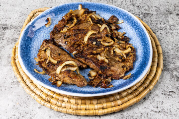 A blue plate of fried pork or beef liver and onions on pieces of toasted bread with knife and fork on a grey table