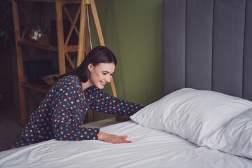 Young woman in pajamas making her bed with a white duvet in a cozy bedroom, enjoying a comfortable...