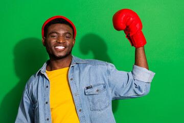 Young stylish man posing with a joyful expression wearing casual denim attire and a red glove against a vibrant green background