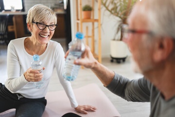 A senior couple sits on yoga mats at home, drinking water and relaxing after a workout session, embracing a healthy and active lifestyle together