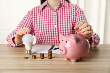 Woman with light bulb putting coin into piggy bank at wooden table, closeup. Energy saving concept
