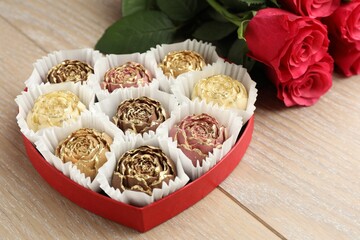 Flower shaped chocolate bonbons in box and red roses on wooden table, closeup