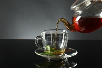 Pouring freshly brewed tea from teapot into cup at dark mirror table, closeup