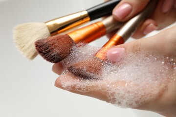 Woman washing makeup brush with soap on light background, closeup