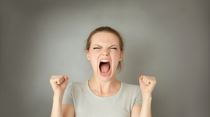 Female model yelling, fists clenched, expressing intense anger during dramatic studio portrait against neutral gray background, conveying powerful emotional conflict