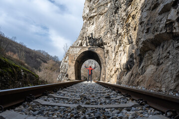 A middle-aged man in a red jacket walks along an old and abandoned railway. A man sits along a railroad track in a rural area, and in front of him large concrete tunnels can be seen.
