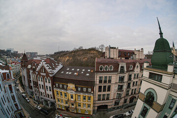 Panoramic cityscape view with mixed architectural styles, two-story buildings some with windows, wet road, overcast sky, muted colors grey, brown, occasional splashes of red and yellow, gives sen © Mister