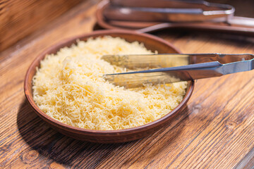 Image of a close-up view of a wooden cutting board with two spatulasserving tongs on it, partially submerged in grated yellow Parmesan-like cheese in a bowl Rustic setting suggested by wooden table