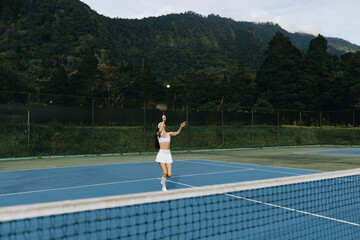 Young female tennis player in white sportswear, serving on outdoor court surrounded by lush green...