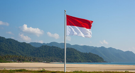The Indonesian flag proudly waves against a bright blue sky, overlooking a beautiful coastline with gentle waves and lush green mountains in the background