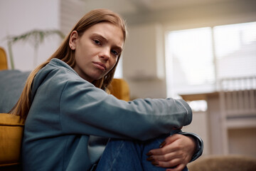 Upset teenage girl hugging her knees while sitting at home looking at camera.