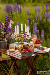 Two glasses of rose wine, flowers in a vase, silverware, fruits. Sunset, golden hour, atmospheric candlelight. Romantic table decor for a loving couple on blooming meadow with purple lupines