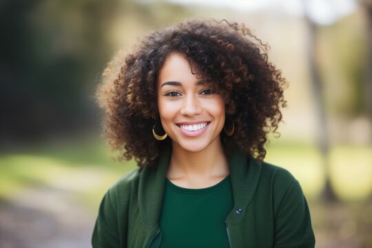 A woman with vibrant curly hair exudes joy while standing in a lush green park. The sunlight bathes her in warmth, creating a cheerful atmosphere. Her smile radiates happiness and confidence